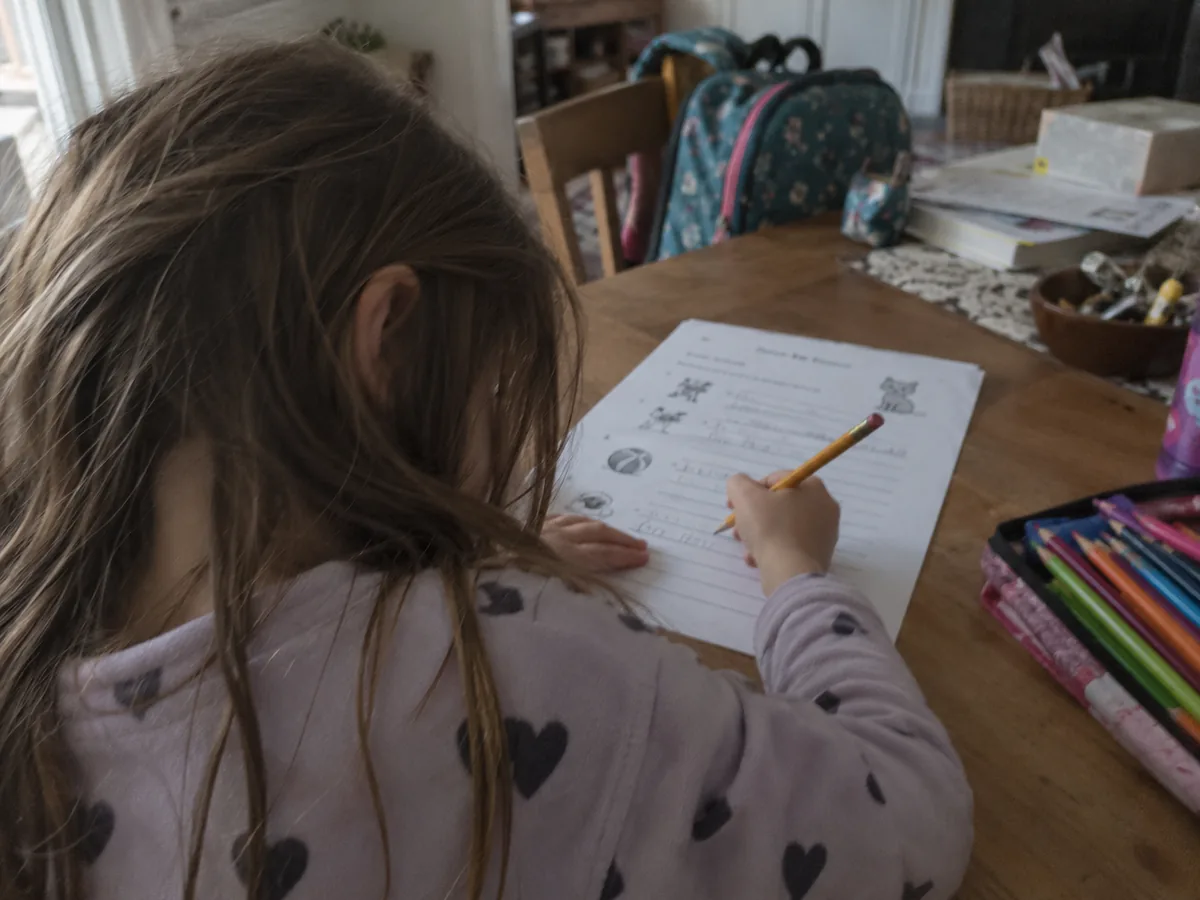 Criança de pijama escrevendo a lápis numa folha impressa, sobre uma mesa de madeira